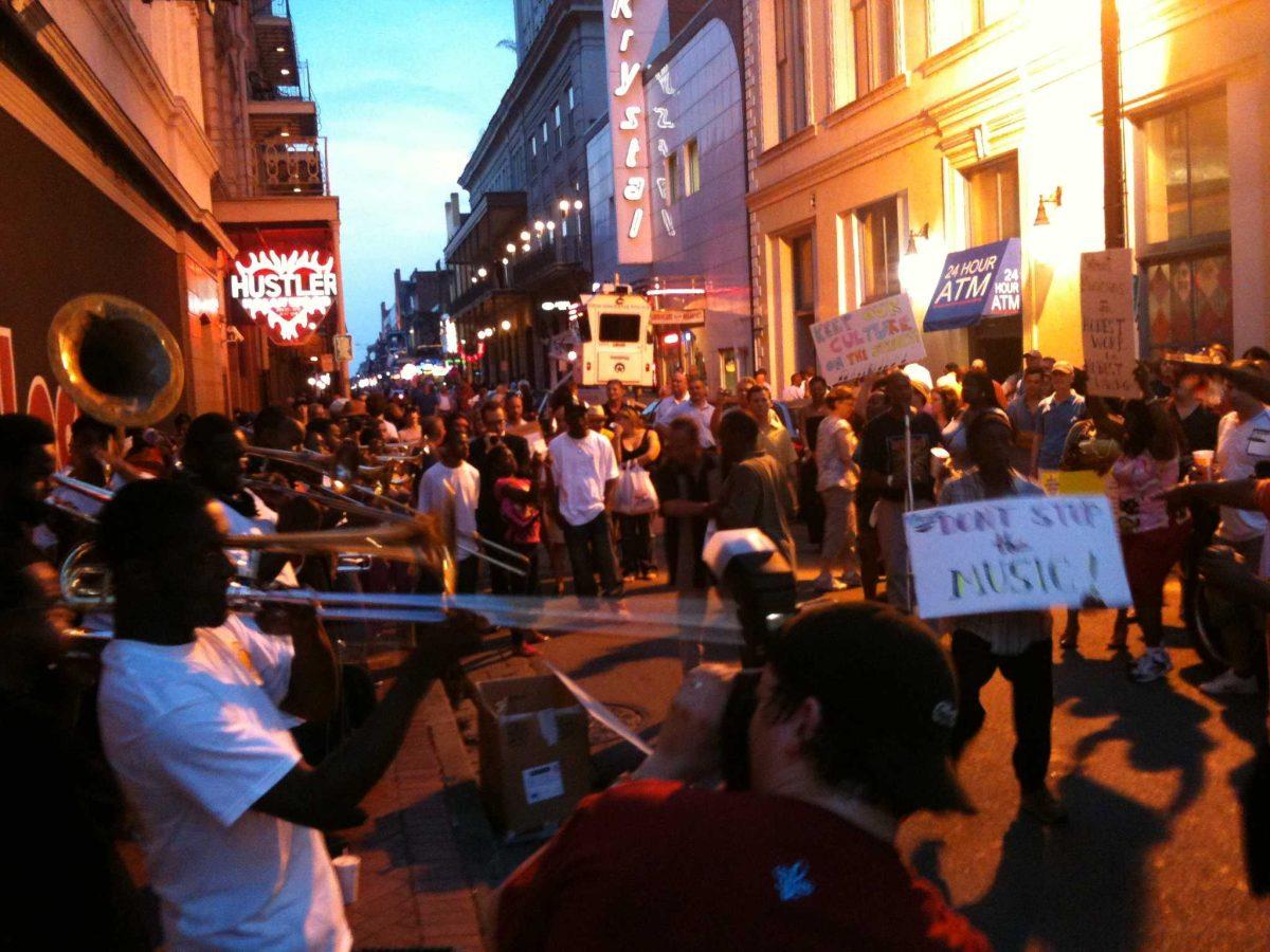 Musicians play on the streets of the French Quarter while onlookers support them with signs.