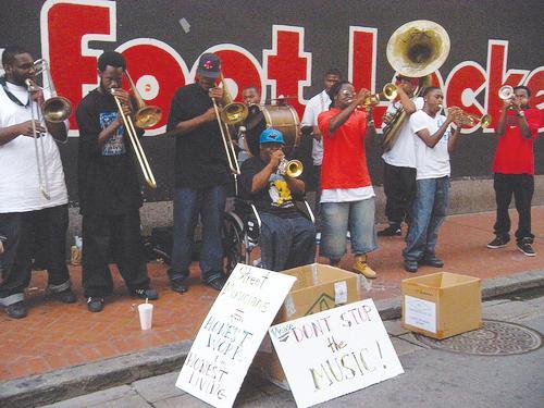 Members of To Be Continued Brass Band play music on a New Orleans street behind a sign reading &#8220;Please Don&#8217;t Stop the Music!&#8221;