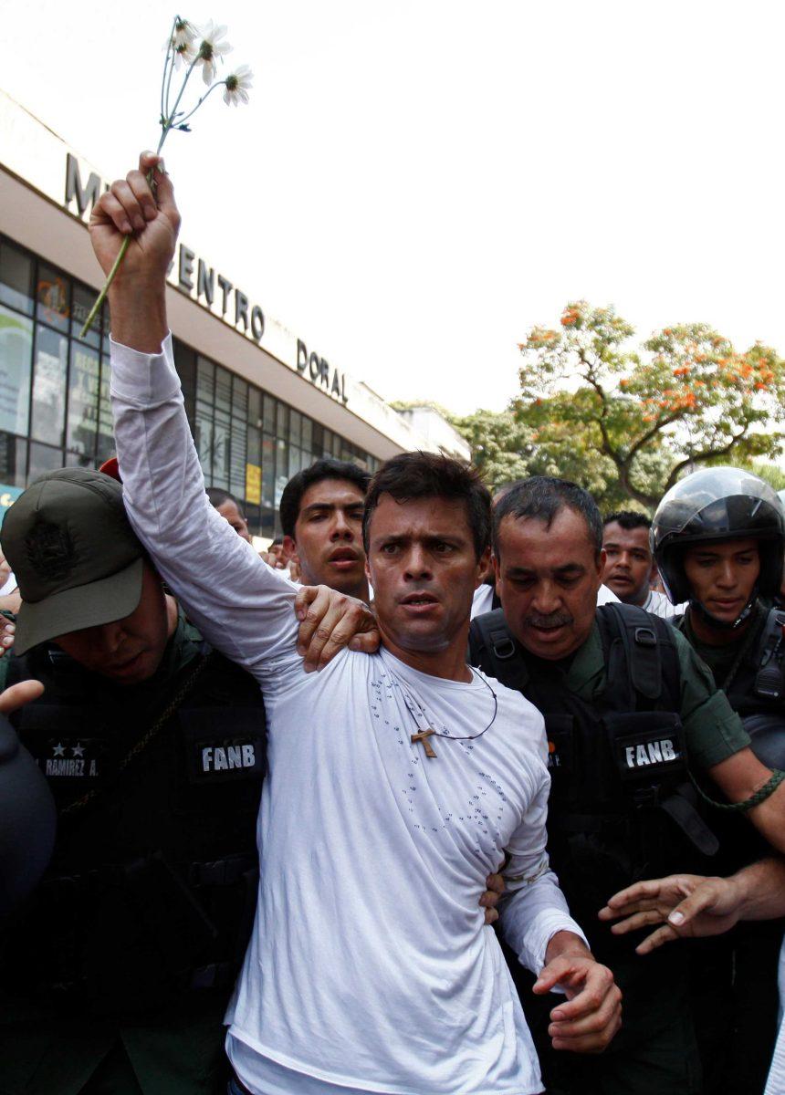 Opposition leader Leopoldo Lopez, dressed in white and holding up a flower stem, is taken into custody by Bolivarian National Guards, in Caracas, Venezuela, Tuesday, Feb 18, 2014. Lopez re-emerged from days of hiding to address an anti-government demonstration and then he turned himself in to authorities Tuesday. Speaking to some 5,000 supporters with a megaphone, Lopez said that he doesn't fear going to jail to defend his beliefs and constitutional right to peacefully protest against President Nicolas Maduro. (AP Photo/Alejandro Cegarra)