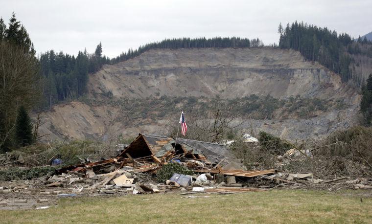 A flag, put up by volunteers helping search the area, stands in the ruins of a home left at the end of a deadly mudslide from the now-barren hillside seen about a mile behind, Tuesday, March 25, 2014, in Oso, Wash. At least 14 people were killed in the 1-square-mile slide that hit in a rural area about 55 miles northeast of Seattle on Saturday. Several people also were critically injured, and homes were destroyed. (AP Photo/Elaine Thompson)