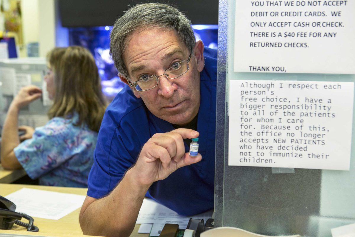 Pediatrician Charles Goodman poses for photo with the MMR vaccine, an immunization vaccine against measles, mumps, and rubella at his practice in Northridge, Calif., Thursday, Jan. 29, 2015. With California gripped by a measles outbreak, Dr. Goodman posted a clear notice on his door and on Facebook: His practice will no longer see children whose parents won't get them vaccinated. At left, is his wife, Judith. (AP Photo/Damian Dovarganes)