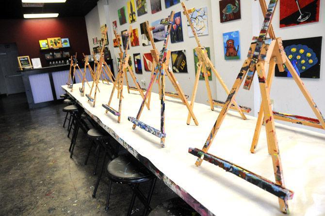 Easels caked with paint stand in front of a wall of completed works at Painting and Pinot on Perkins Road.