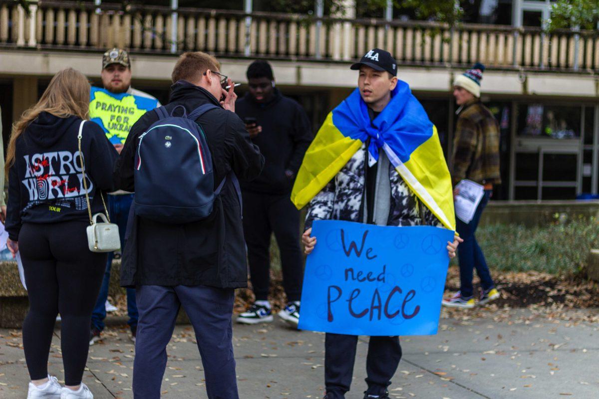 A Kazakh student stands Friday, Feb. 25, 2022, with the Ukraine flag wrapped around his shoulders and a sign saying "We need Peace" in Free Speech Plaza.