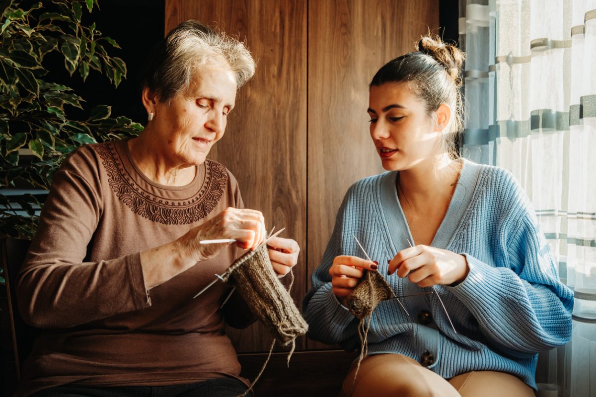Senior caucasian woman teach her granddaughter how to knit with needles sit by the window during the day